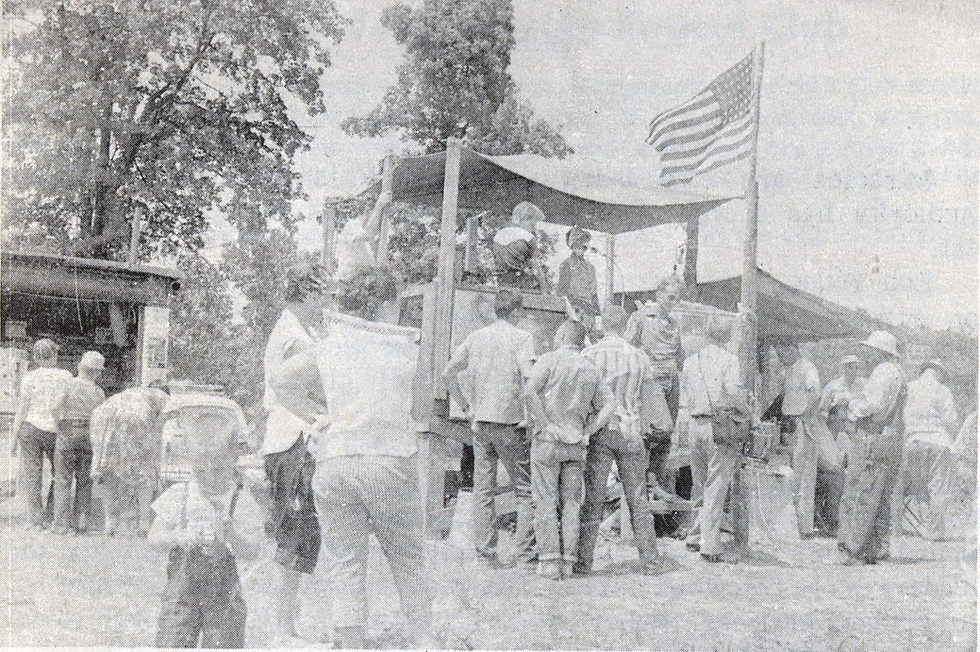 A crowd of people standing outside around a raised, covered wooden platform look at a speaker in front of a microphone. An American flag waves next to the platform. 