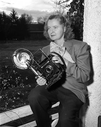 A black and white photo of a woman witting on a front porch railing and holding an old fashioned camera with a large flash bulb 