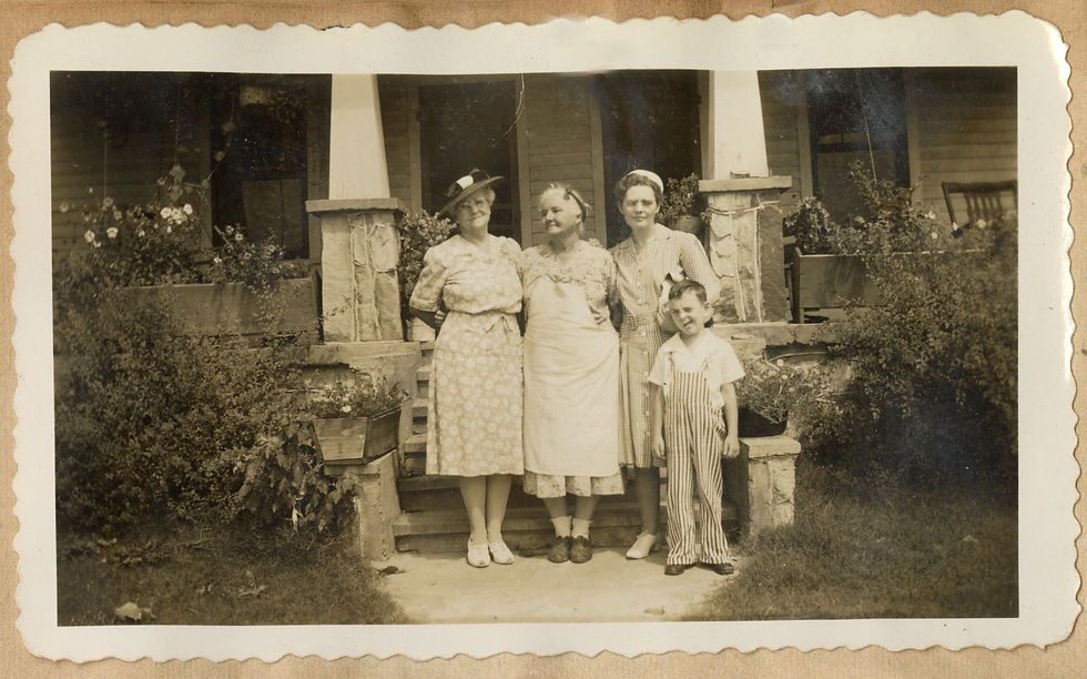 Three women stand side by side in front of a house's stone craftsman-style front porch. There is a young boy in striped overalls winking at the camera and standing in front of one of the women. 