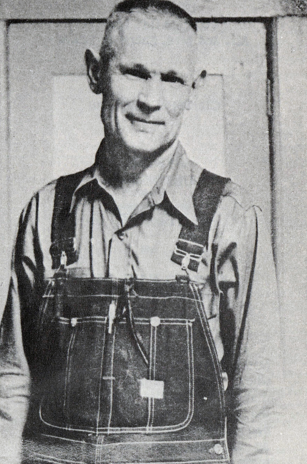 A man with short-cropped hair and a heavy brow wears overalls and squints into the camera