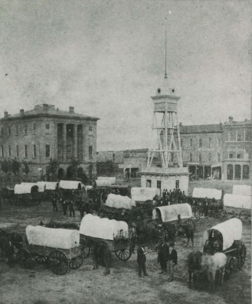 A black and white photo of a dozen covered wagons gathered around a monument in the center of the square. People and horses stand about. A stone building with large columns stands in the background to the left. 