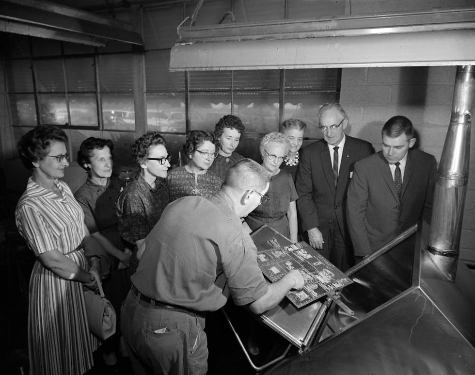 A group of women gather around a man standing by a machine holding an engraving