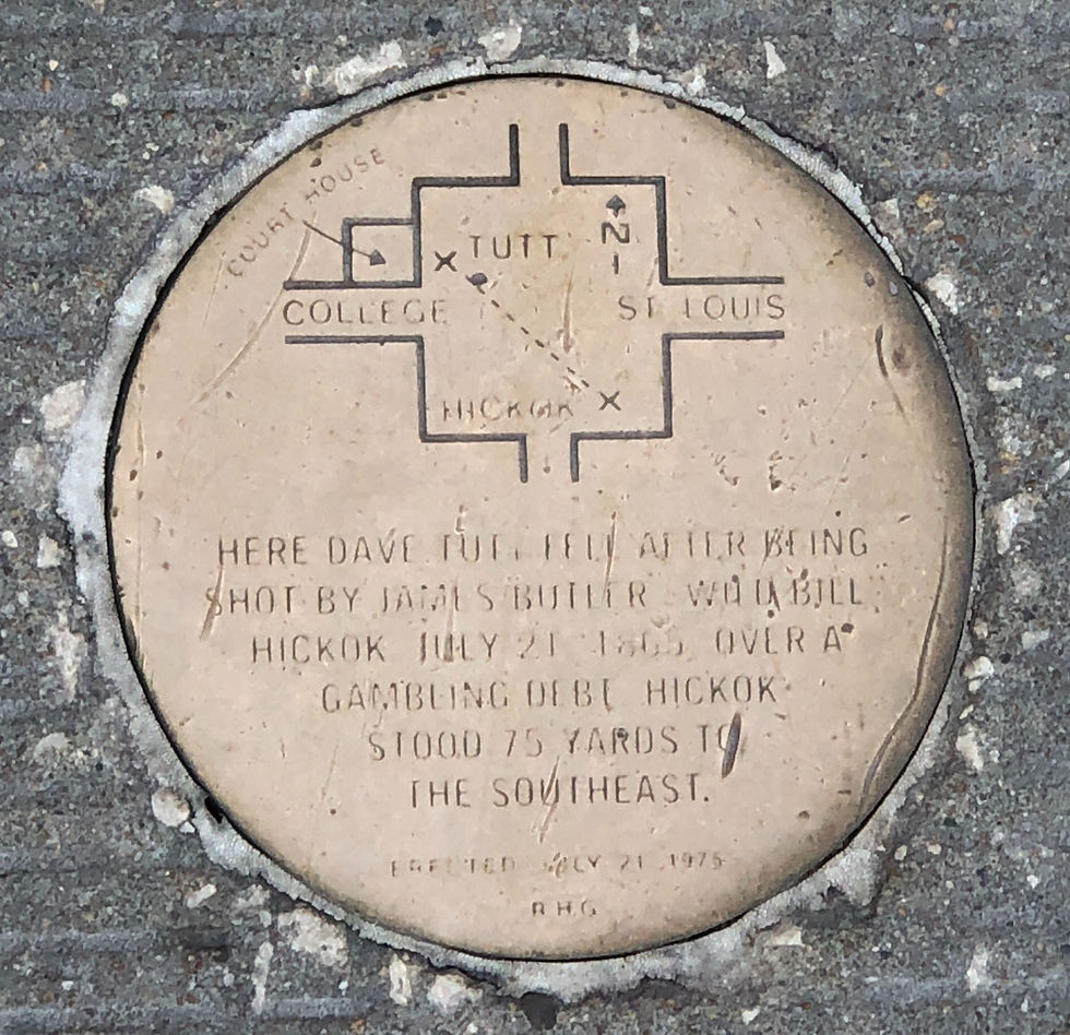 A shiny brass round medallion embedded in asphalt with a map of the Springfield square etched in it. The map shows the locations of Tutt and Hickok along with an inscription about Tutt's death. 