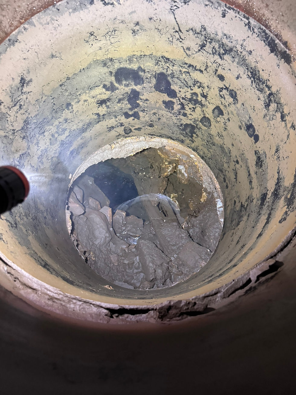 a flashlight illuminates a corrugated metal culvert, at the bottom of which stands mud, water, and a peek into a deep cavern below the water's surface