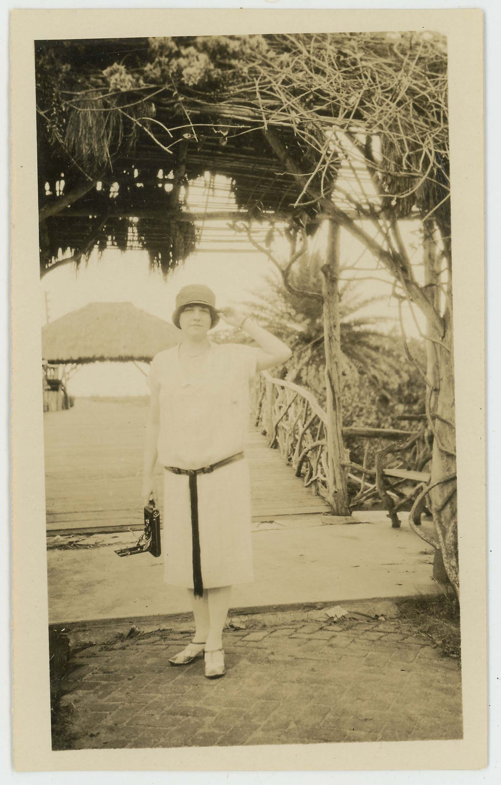 A woman wearing a bucket hat and holding a camera stands at the foot of a dock lined with tropical trees and a thatched roof hut.