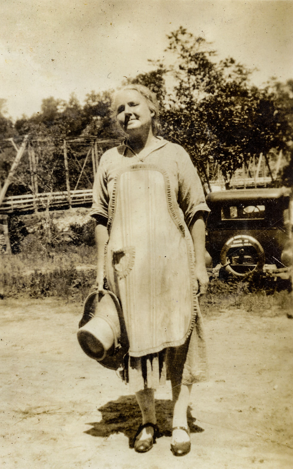 A sepia-toned image of a woman in a simple working dress, holding a hat in her right hand, and standing in front of a parked Model T style vehicle. 