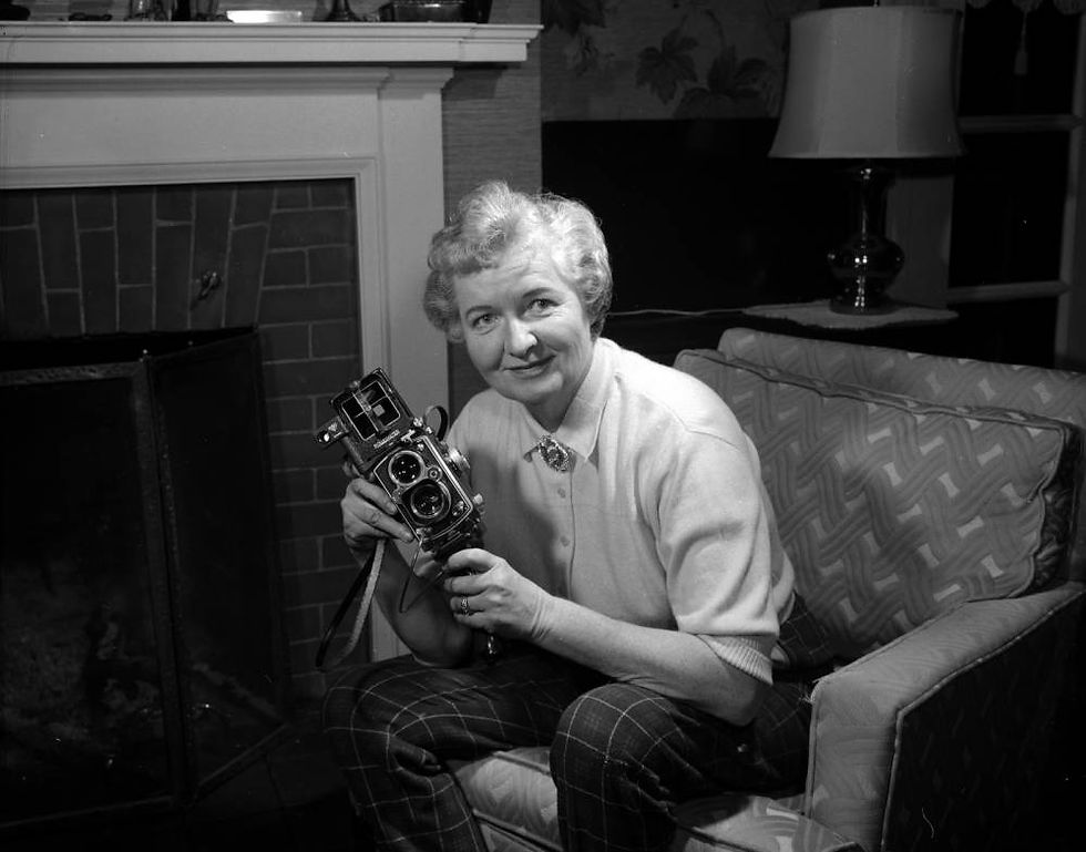 A woman sits in an armchair in front of a hearth while holding an old-fashioned handheld camera