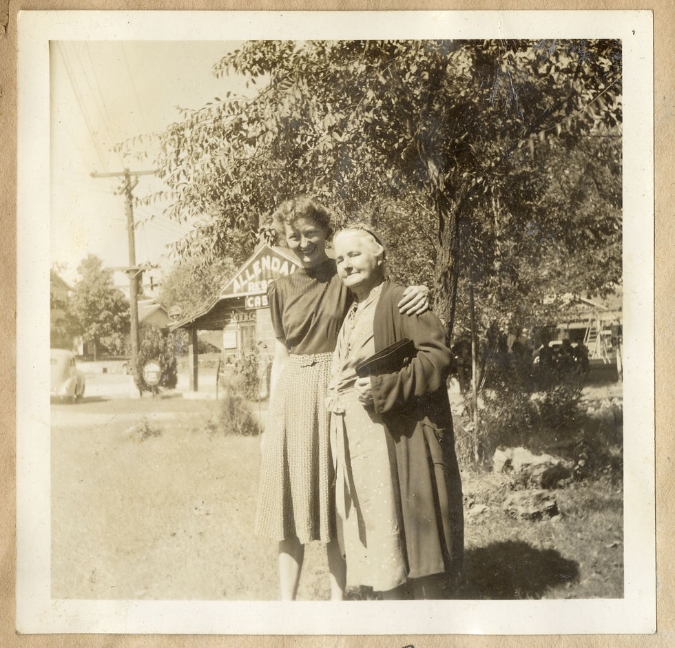 A tall woman in a blouse and textured skirt wraps her left arm around the shoulders of an older woman wearing a long coat and cotton dress, holding a binder or folder. They are standing outside in front of a log cabin labeled "Allendale Resort Cabin Office"