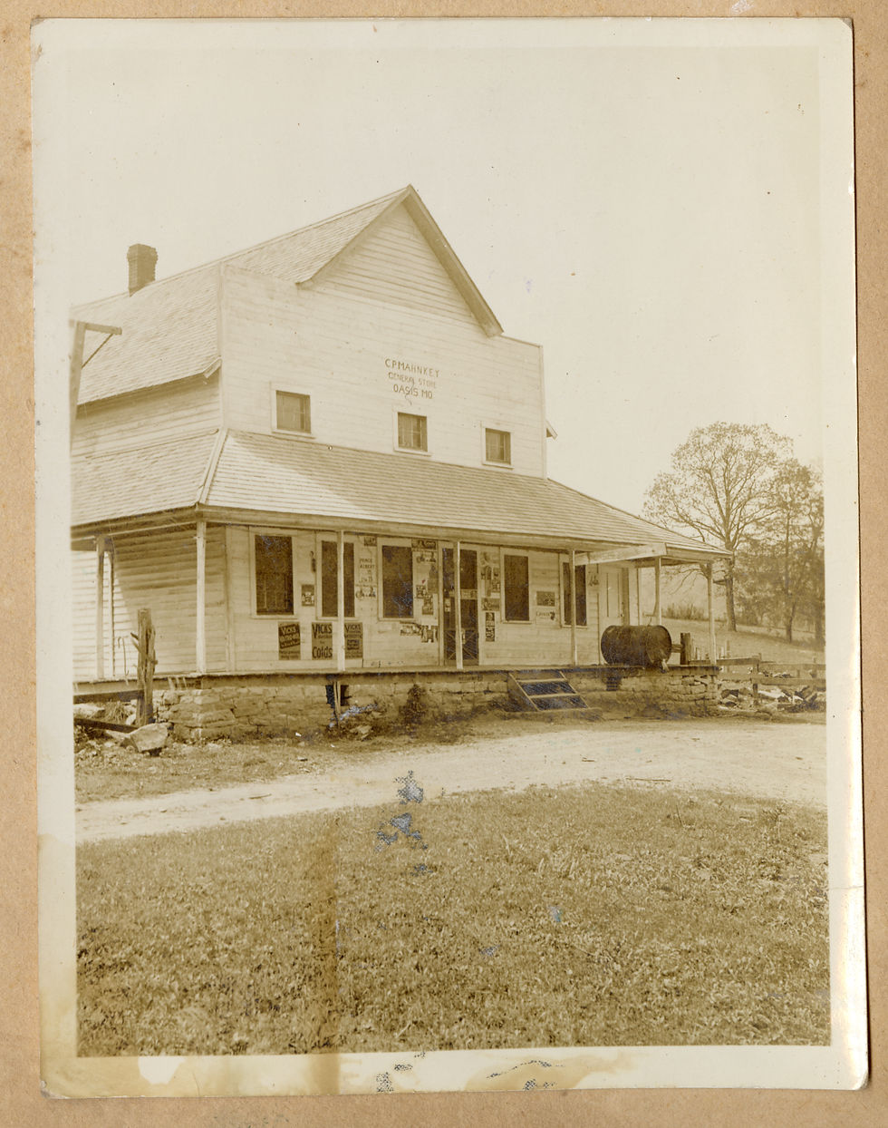 An exterior view of a two story building with a wide front porch and wooden siding. A gravel drivewaylines the front, and various signs are plastered on the front of the building. A sign on the second story says "C.P. Mahnkey General Store Oasis, Mo." 