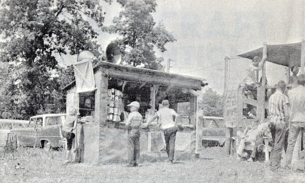 Young men stand in front of a concessions or vendors stand made of wood and canvas in an outdoor field. Large loudspeakers are on top of the stand, and parked cars are visible in the background.