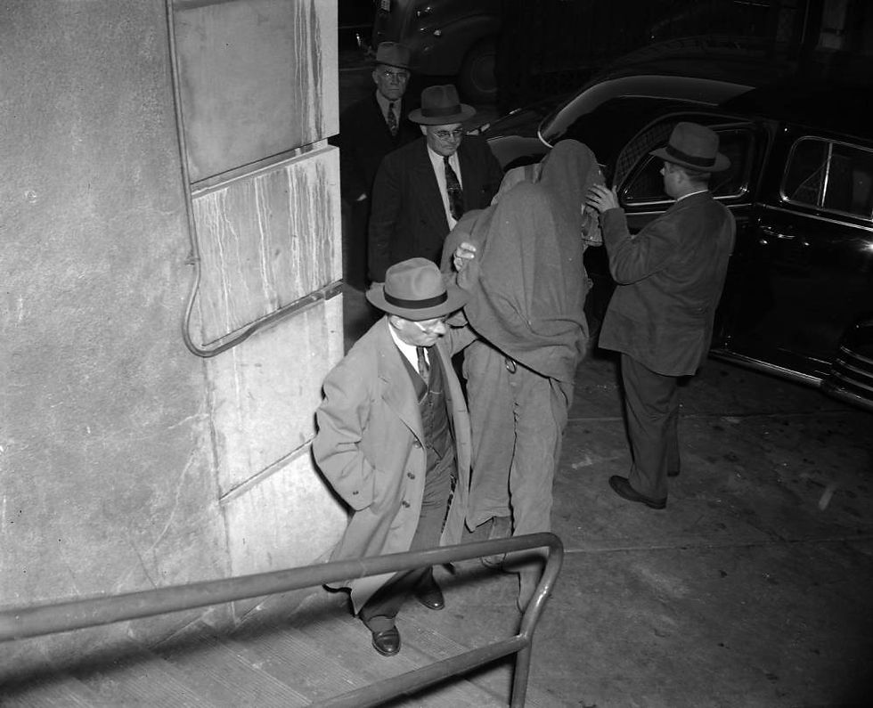 Men in suits lead two men with their heads covered in a blanket up a flight of stairs 