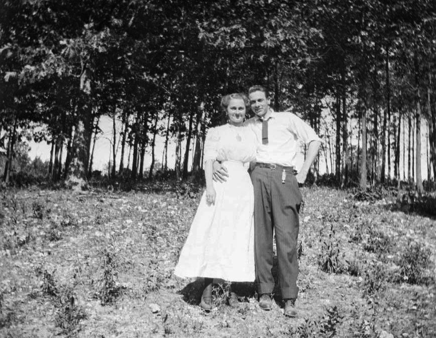 A young couple stand in a clearing in front of trees. A young man wearing a tie and striped trousers has his right arm around the waist of a young woman wearing a short sleeved blouse and long skirt.