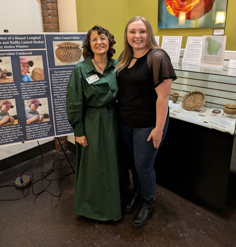A woman wearing a green dress and glasses stands next to a woman in a black top and jeans. They are posing next to an instructional poster and a display case of woven artifacts