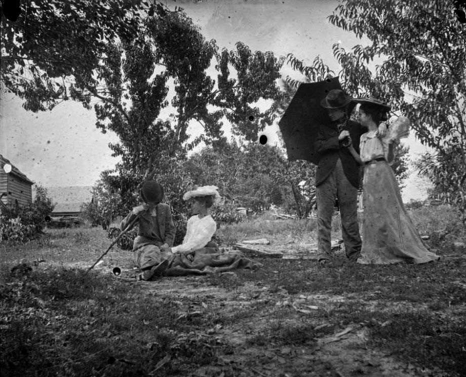 Two couples outside near trees and a house or barn in the background. One couple sits on the ground, the man holding a walking stick and the woman wearing a hat. Another couple stands nearby looking on. The man is holding an umbrella with his arm draped over his partner's shoulder. The woman is holding a fan.