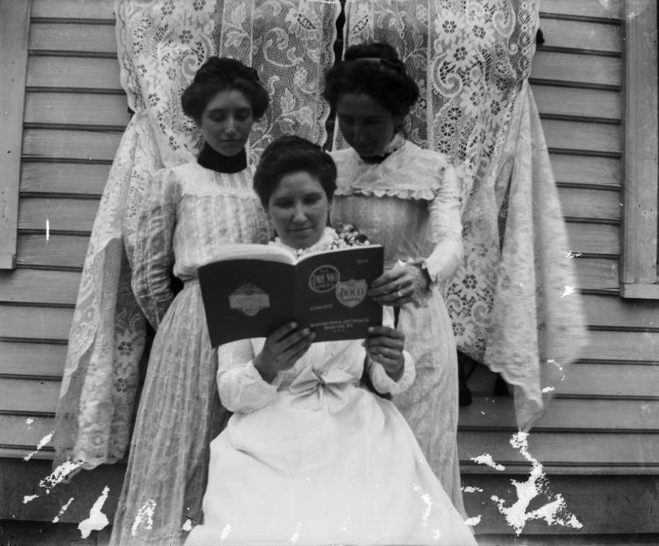 Three women stand in front of a house with siding and lace curtains. Two women stand behind a seated woman. All three are looking at a catalog or book and are wearing long dresses.