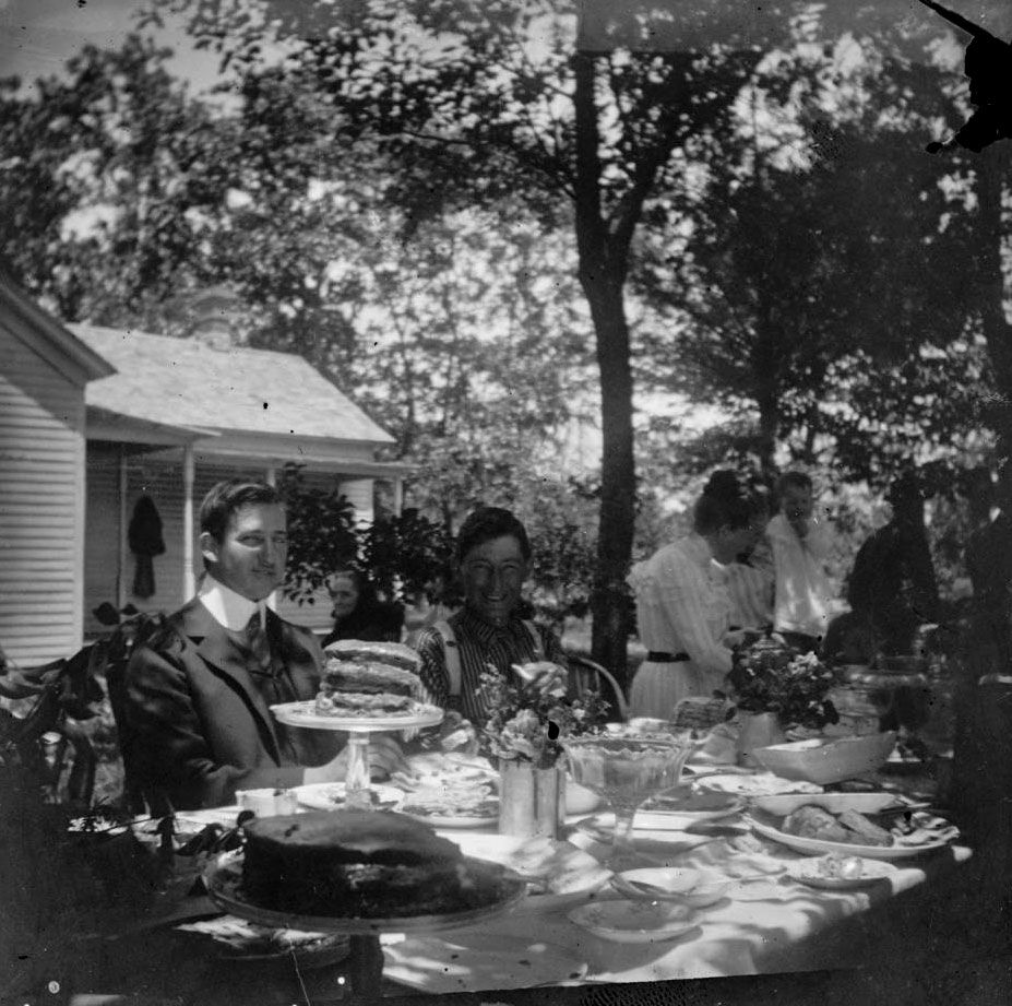 A family gathers at a table outdoors adorned with flowers, cake plates, and dishes. Trees, plants, and family members surround the table. Two young men are seated side by side in focus looking at the camera.