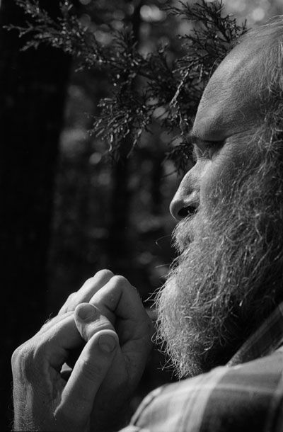 A closeup of a bearded man with his hands clasped. A tree limb behind his head is also in focus.