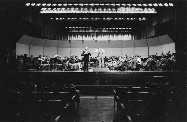 A wide shot of an orchestra on a dramatically lit stage. A conductor stands in front of the orchestra, and a man in a black suit stands to the left of the conductor as he plays his horn. The silhouettes of audience members can be seen in the front row.
