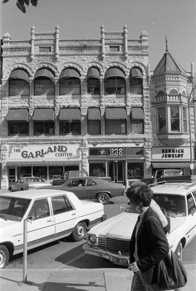 Cars parked on a busy street in front of an ornate building with signs that read "The Garland Center", "328", and "Rennick Jewelry". A woman and another person walk in front of the camera.
