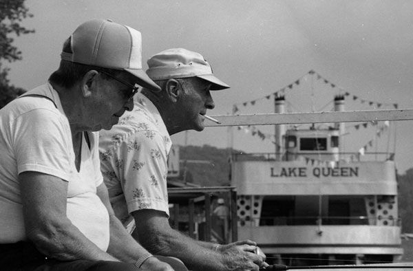 Two older men sit side by side holding fishing lines over the edge of a doc. Both men are wearing caps, and one man has a cigarette dangling out of his mouth. A boat titled "The Lake Queen" can be seen in the background.